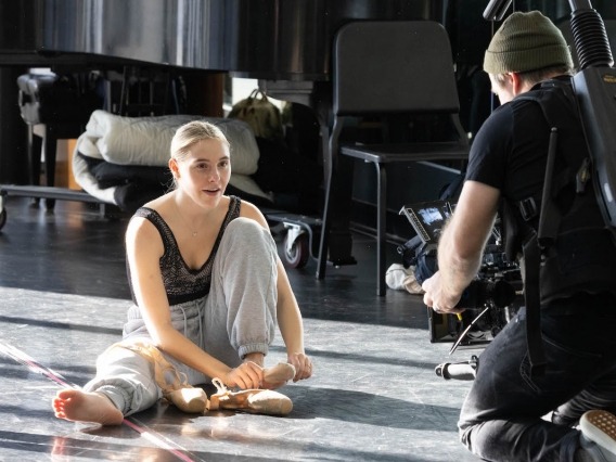 a student dancer stretches on the floor while a man in a black shirt films her