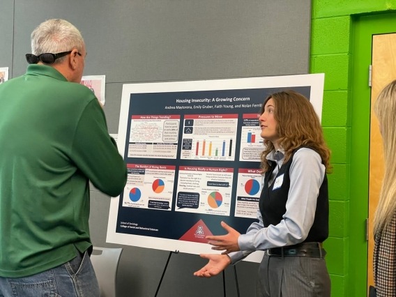 A person wearing a long sleeve shirt, grey slacks and a black vest stands in front of a chart with graphs and data while speaking to a man in a green long sleeve shirt.