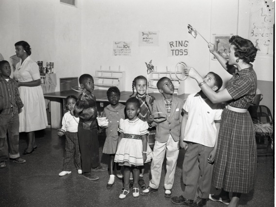 A black-and-white picture from the mid century depicting a group of young children listen to a woman giving directions for a ring toss game.