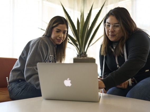 Two students sitting at a table looking at a laptop computer.